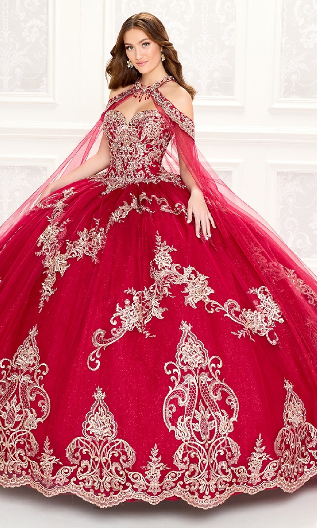 A young woman wears the Quinceanera Dress PR30086 By Princesa, featuring red fabric with gold embroidery, off-the-shoulder straps, a full skirt, and a sheer cape, as she stands before a white decorated wall.